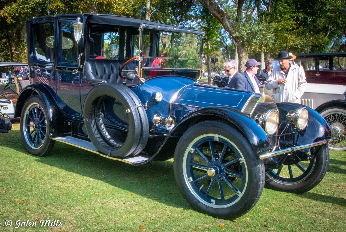 A classic vintage automobile with blue bodywork and large spoke wheels displayed in an outdoor setting with people admiring it.