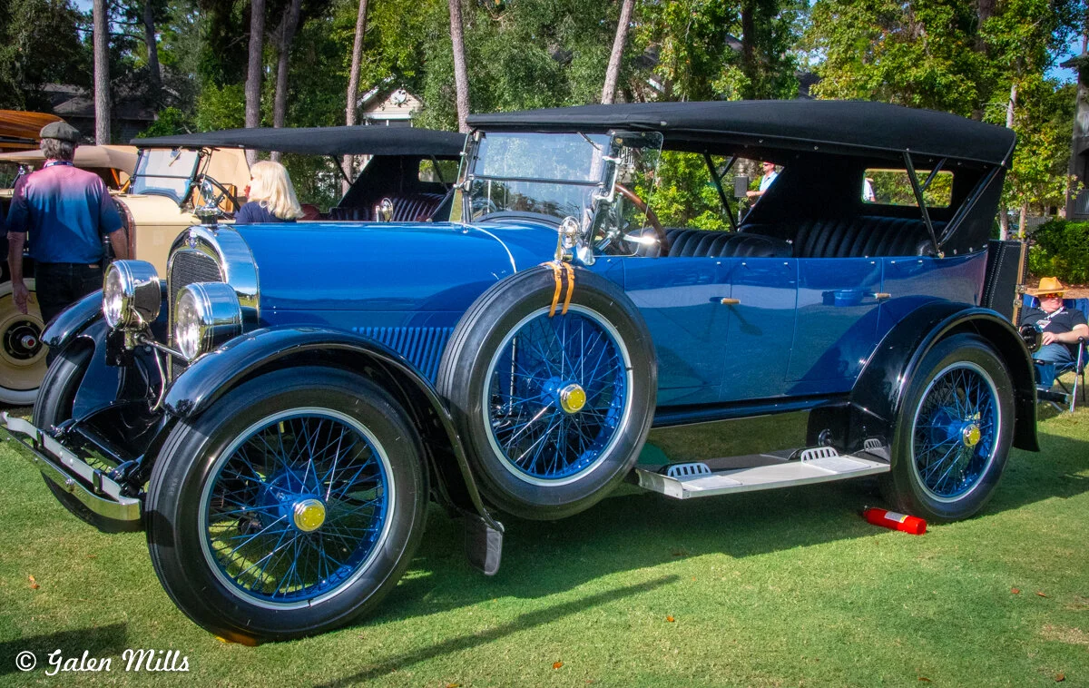 Vintage blue convertible car with black soft top and wire-spoke wheels, displayed outdoors with trees and people in the background.