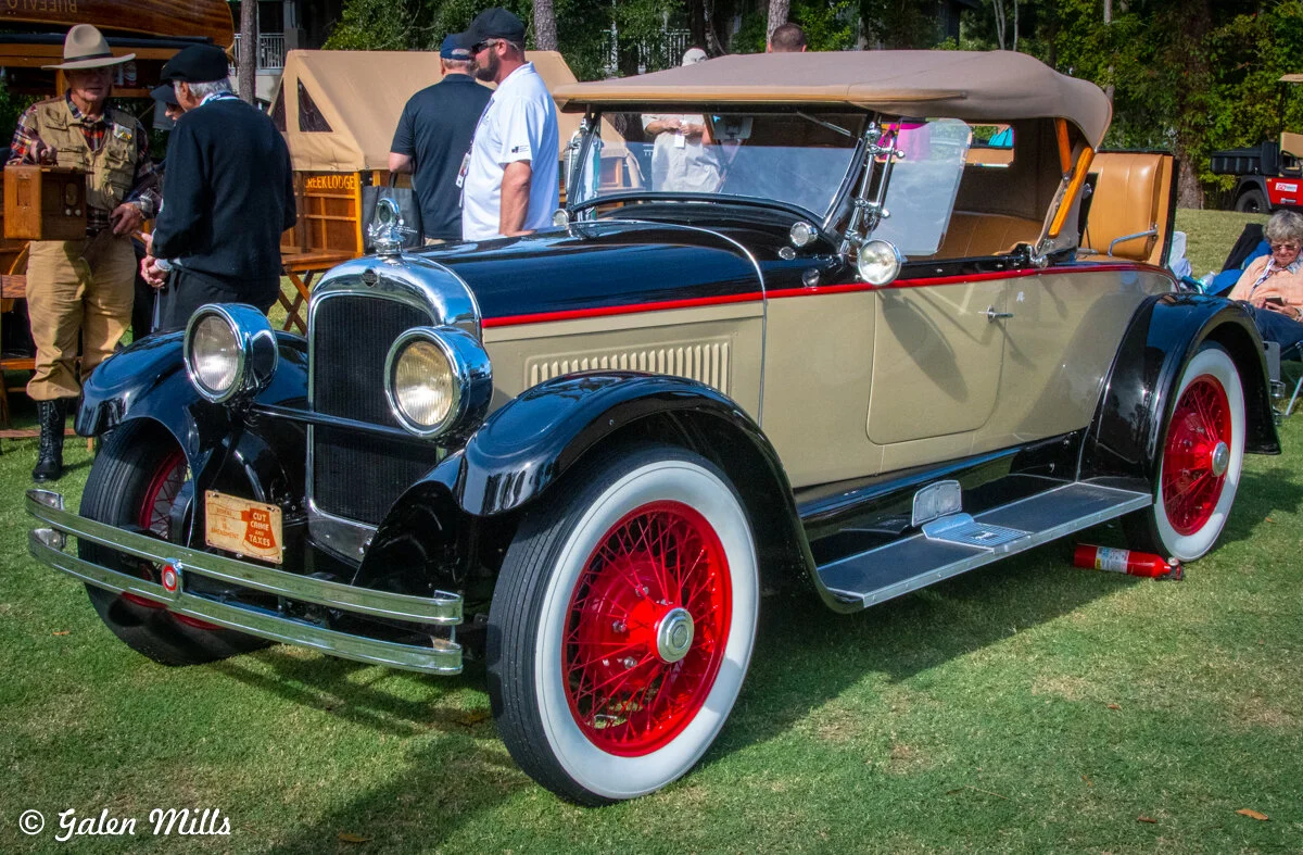 Vintage classic car with red wire wheels and beige body, parked on grass at an outdoor event with people nearby.