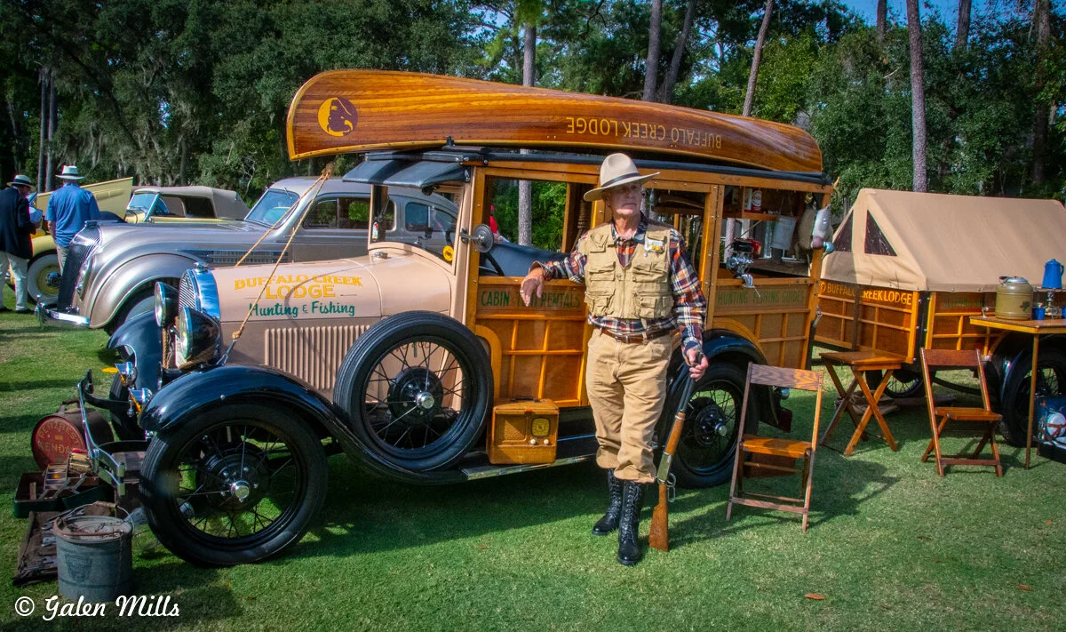 Vintage hunting and fishing vehicle with 'Buffalo Creek Lodge' branding, featuring a canoe on top, showcased at an outdoor event. A man in hunting attire stands beside it, and camping gear is displayed nearby.
