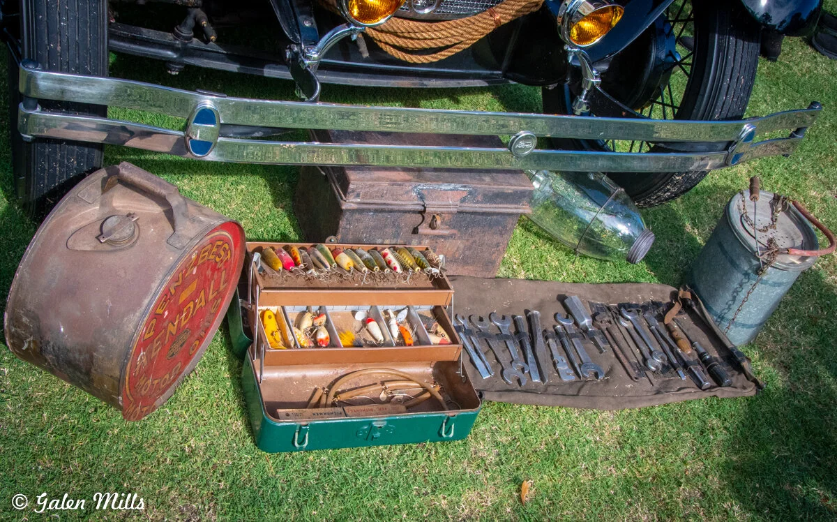 Vintage car bumper and accessories, including a large oil can, a toolbox with colorful bobbers, wrenches, and a metal container on green grass.