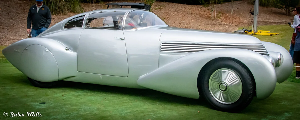 A sleek, silver vintage car with a streamlined design on display outdoors.