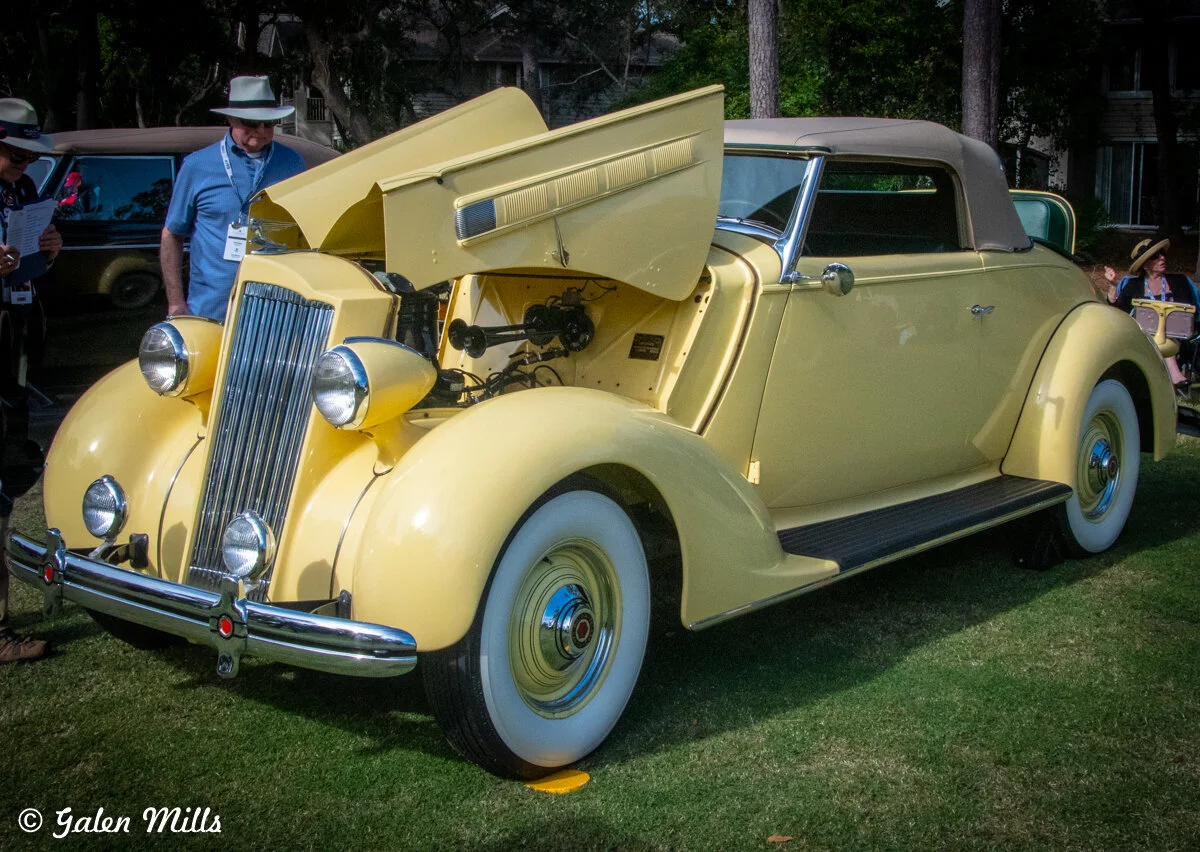 A vintage yellow convertible car with its hood open, showcasing the engine. The car is displayed outdoors on grass, surrounded by people observing.