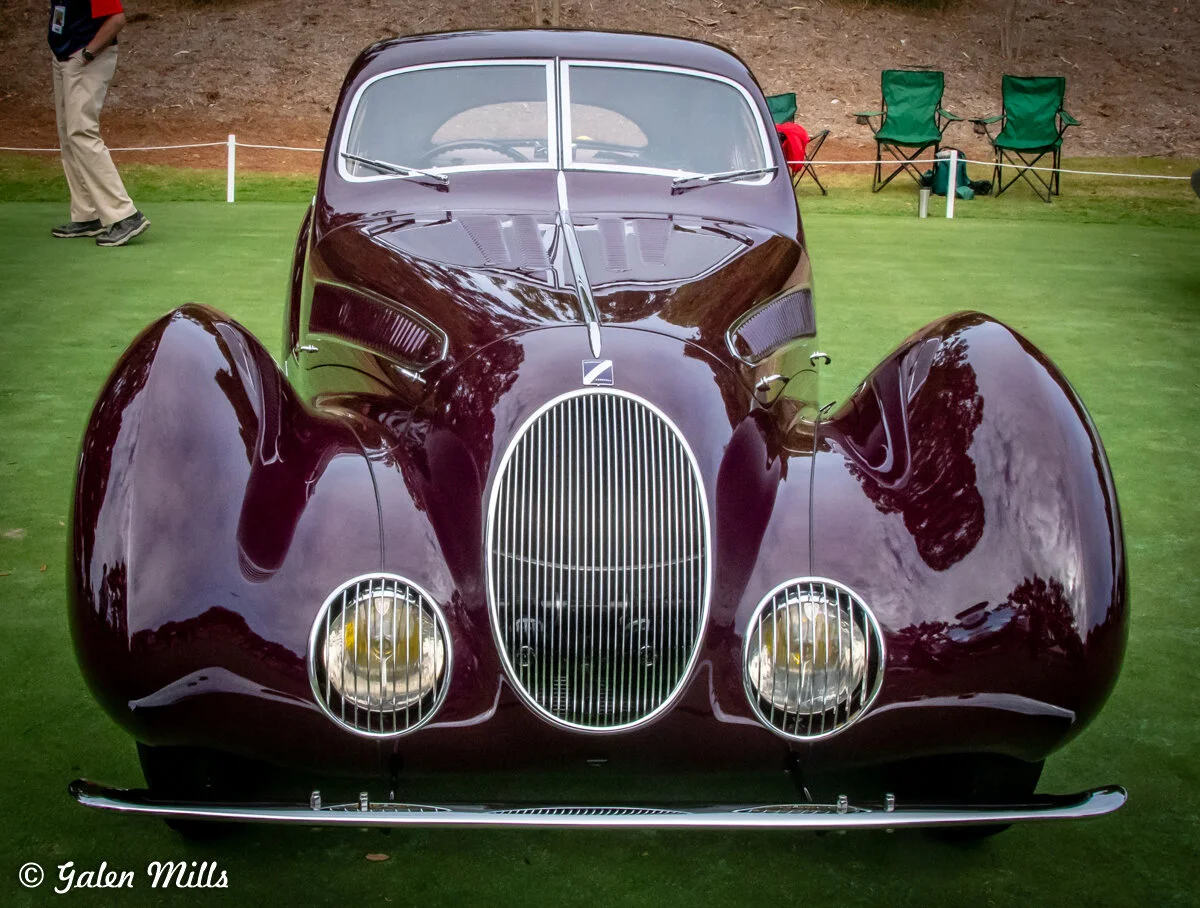 Vintage maroon classic car with distinctive rounded shapes and large headlights displayed on green grass with chairs and a person in the background.