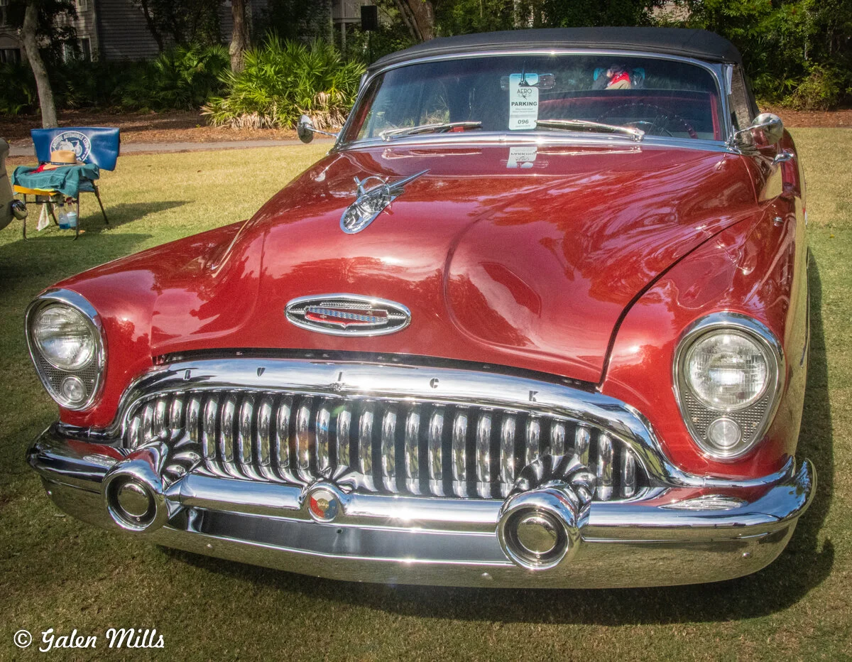 Vintage red Buick car with chrome details on display at a car show. Front view showing grill and headlights.