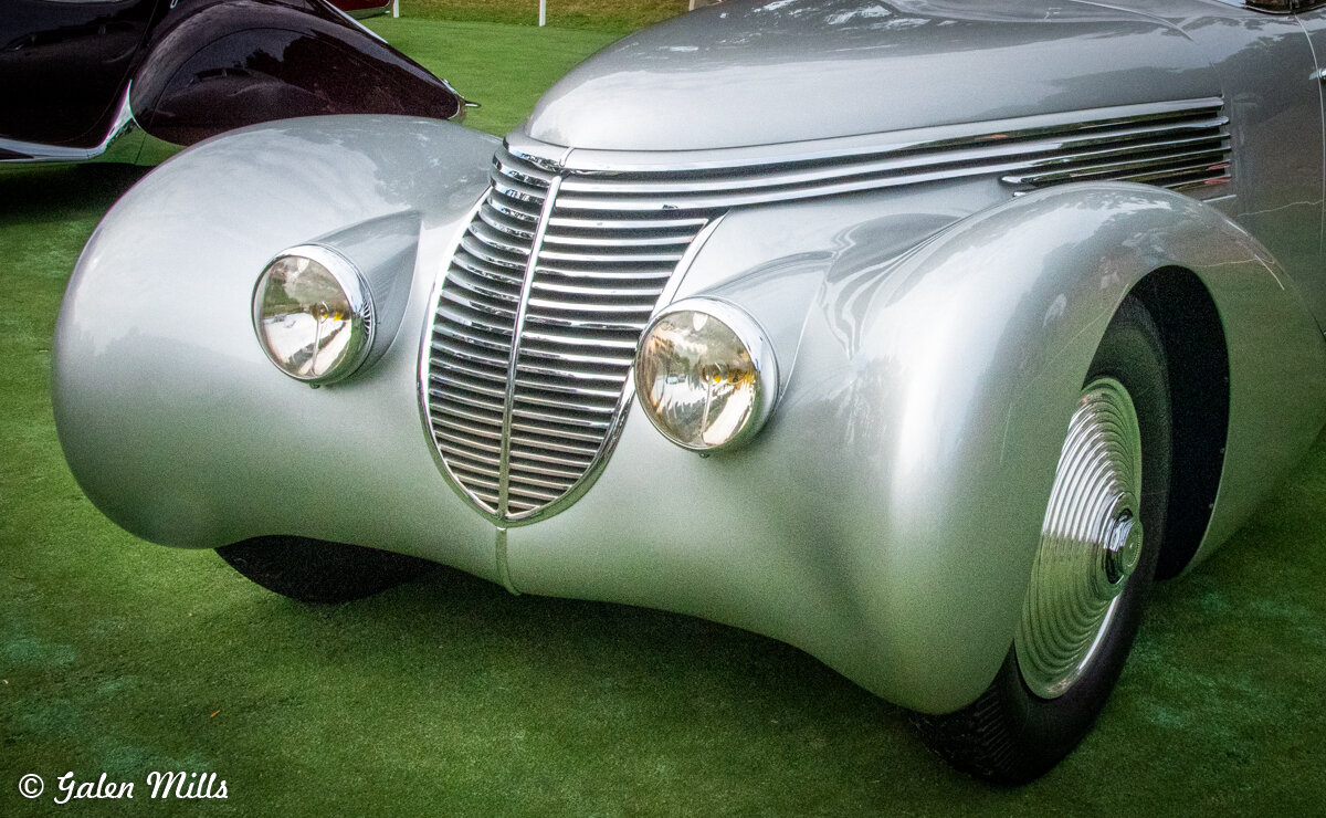 Close-up of a vintage silver car with prominent headlights and a distinctive grill design on a grassy surface.