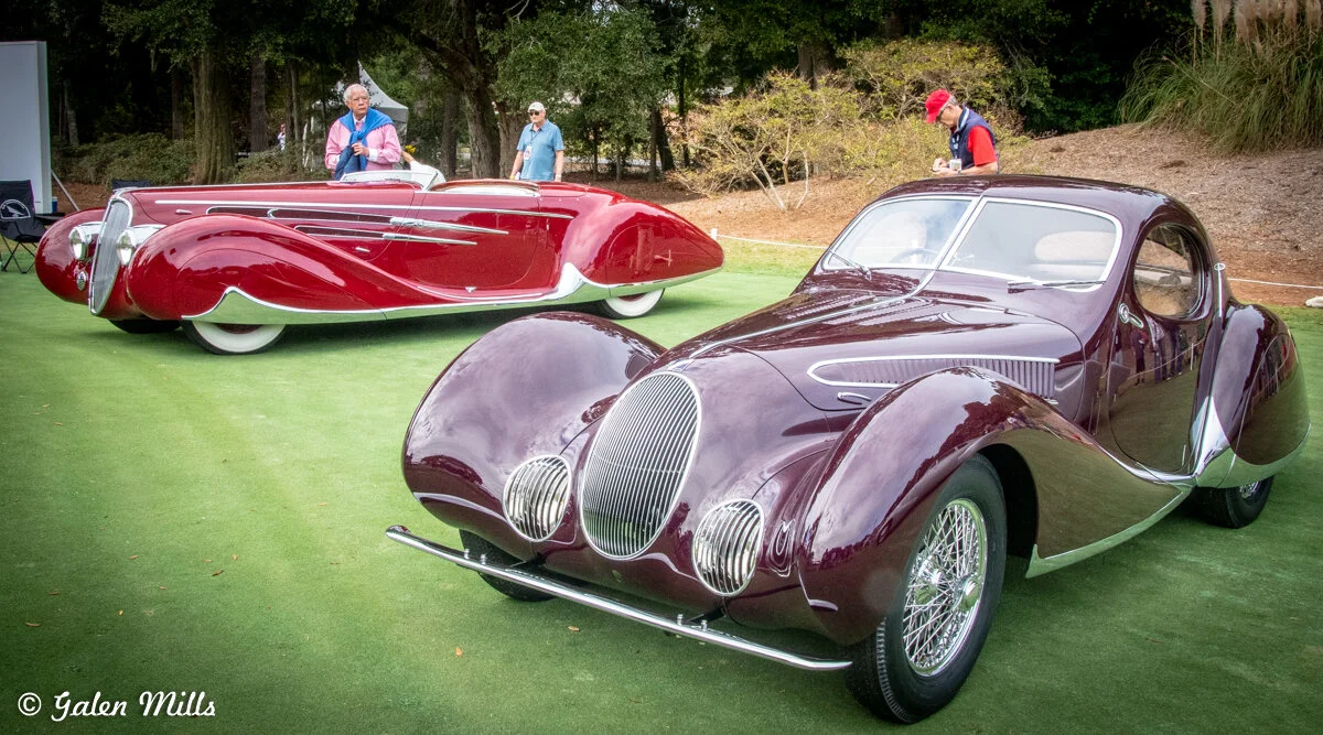 Two vintage luxury cars displayed on a grassy area, with a red convertible in the background and a burgundy coupe in the foreground. Several people stand nearby observing the cars.