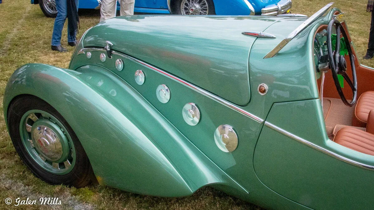 Close-up of a vintage green convertible car with round chrome details, brown leather seats, and a black steering wheel, parked on grass.