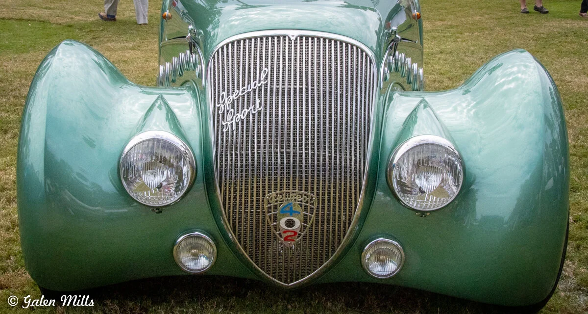 Front view of a green vintage car with prominent headlights and a chrome grille, featuring classic design elements typical of pre-war automobiles. The car is displayed on grass.