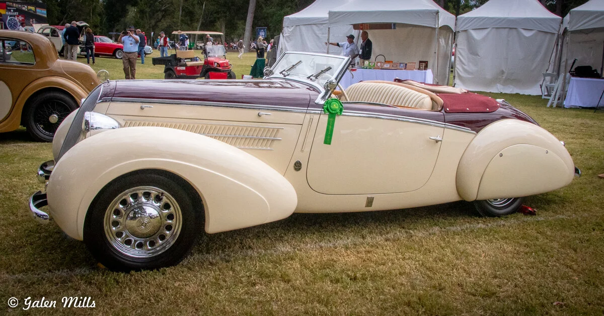 Vintage cream and burgundy convertible car with a ribbon on the door, displayed at a car show with tents and people in the background.