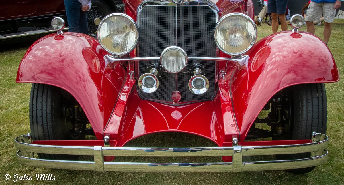 Front view of a red vintage car with large headlights and chrome details, displayed at a car show.