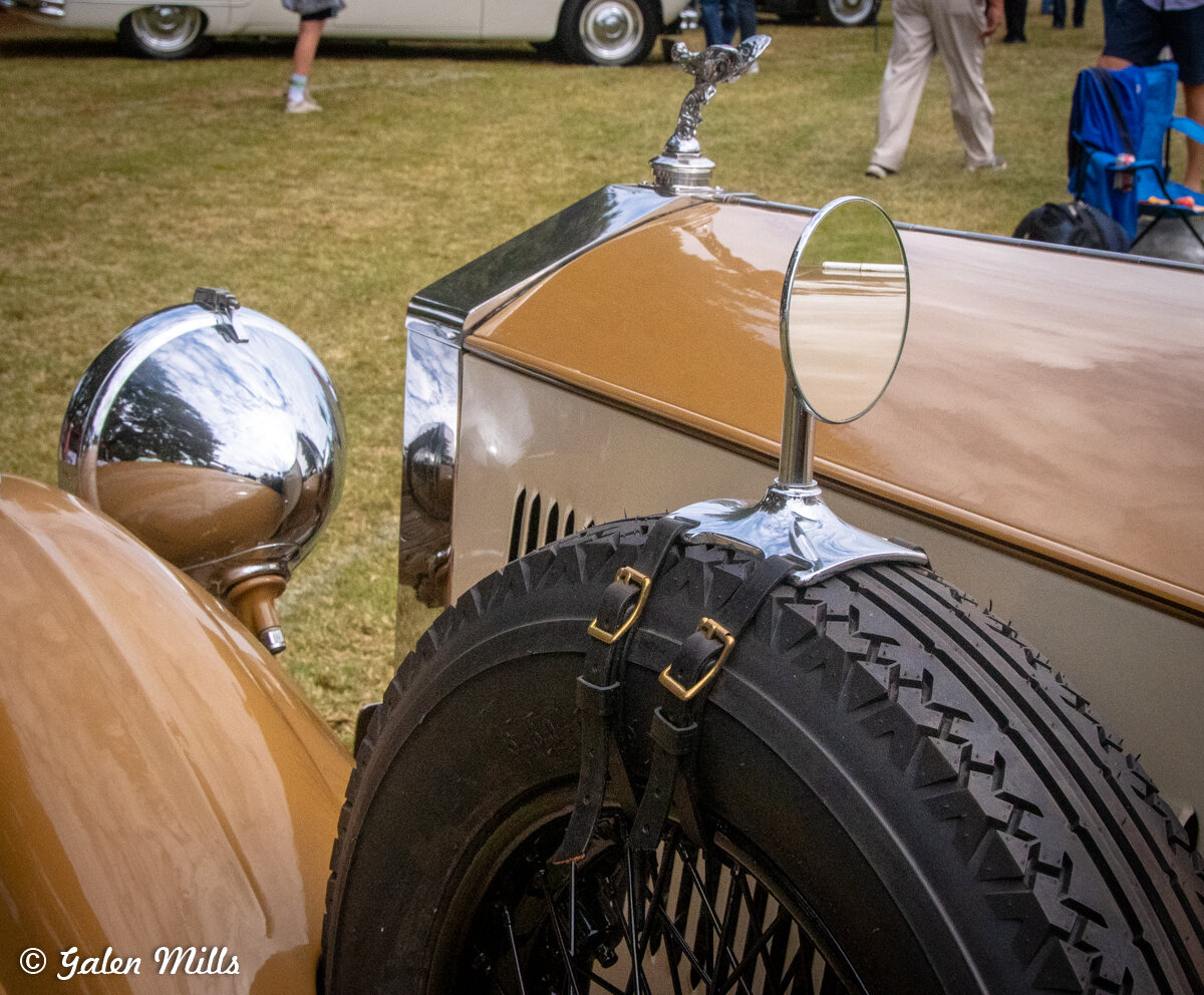 Close-up of a vintage car with a side mirror and spare tire, featuring a detailed hood ornament and fender.