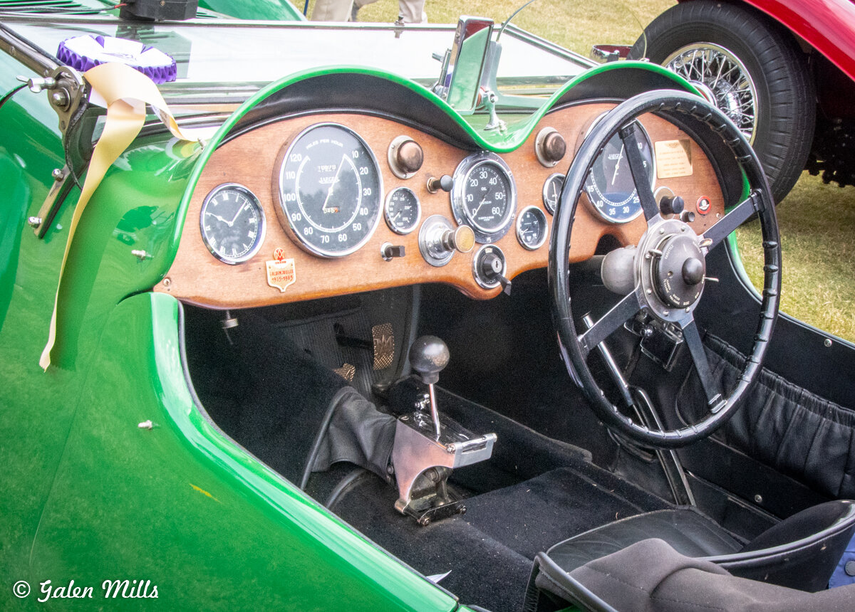 Vintage car interior with wooden dashboard and classic steering wheel, featuring analog gauges and a manual gear shifter, in a green vehicle with visible ribbon.