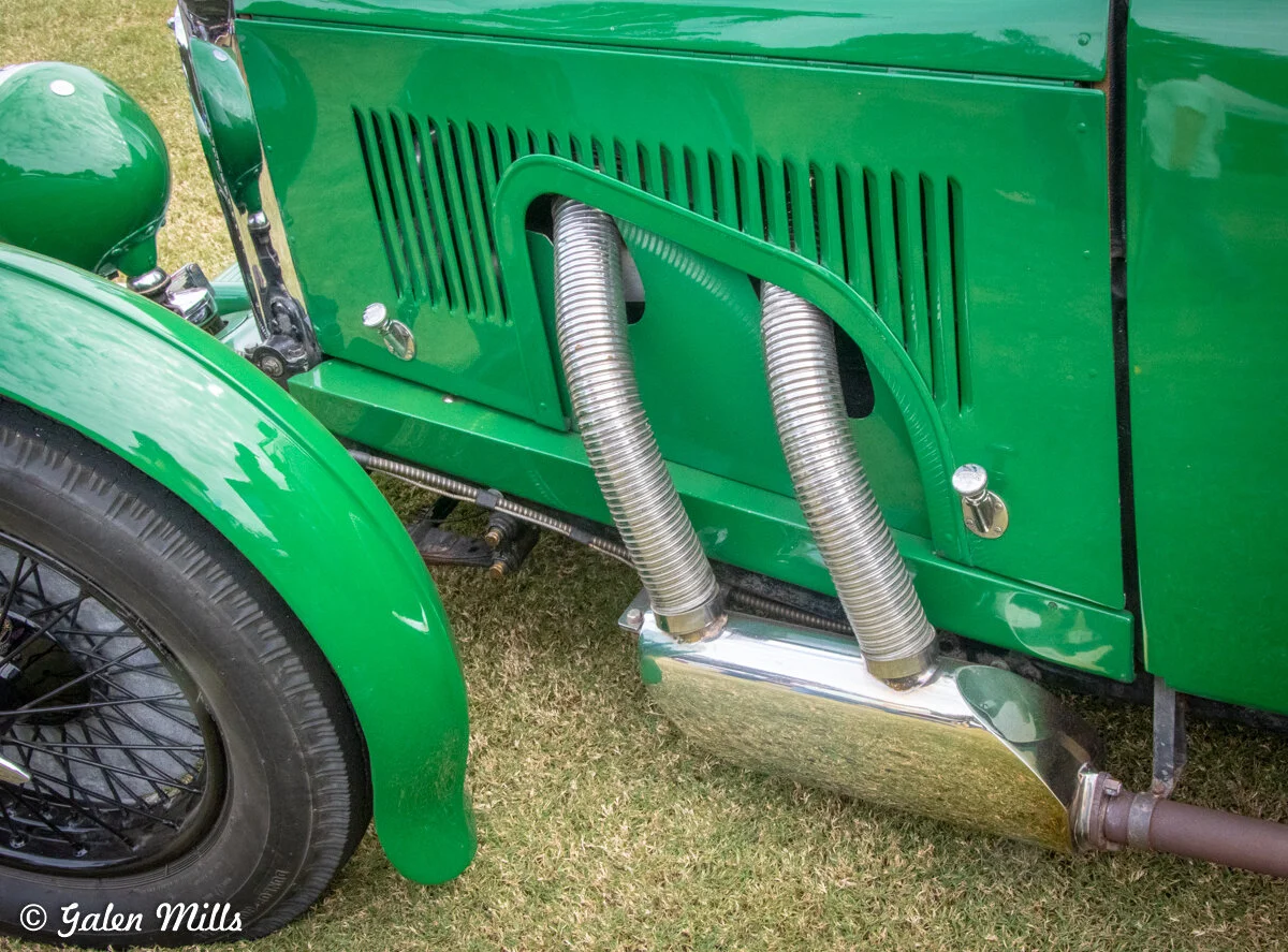 Close-up of a green vintage car's side, featuring chrome exhaust pipes and a black spoked wheel on grass.