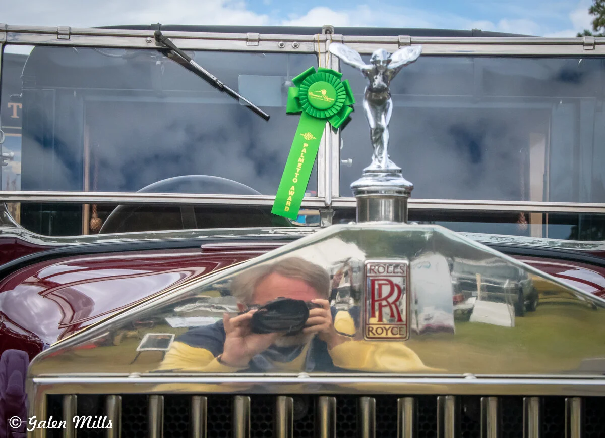 Front view of a Rolls Royce car with a green award ribbon, featuring a hood ornament and reflection of a person taking a photo.