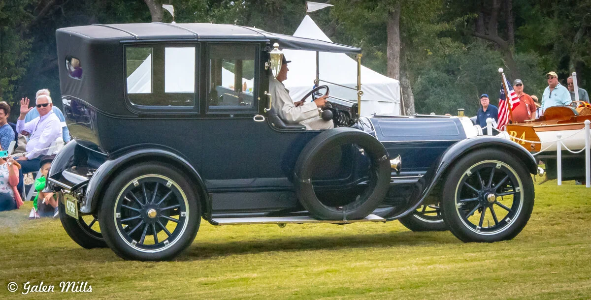 Vintage car on grass with a driver, surrounded by onlookers and a boat in the background.