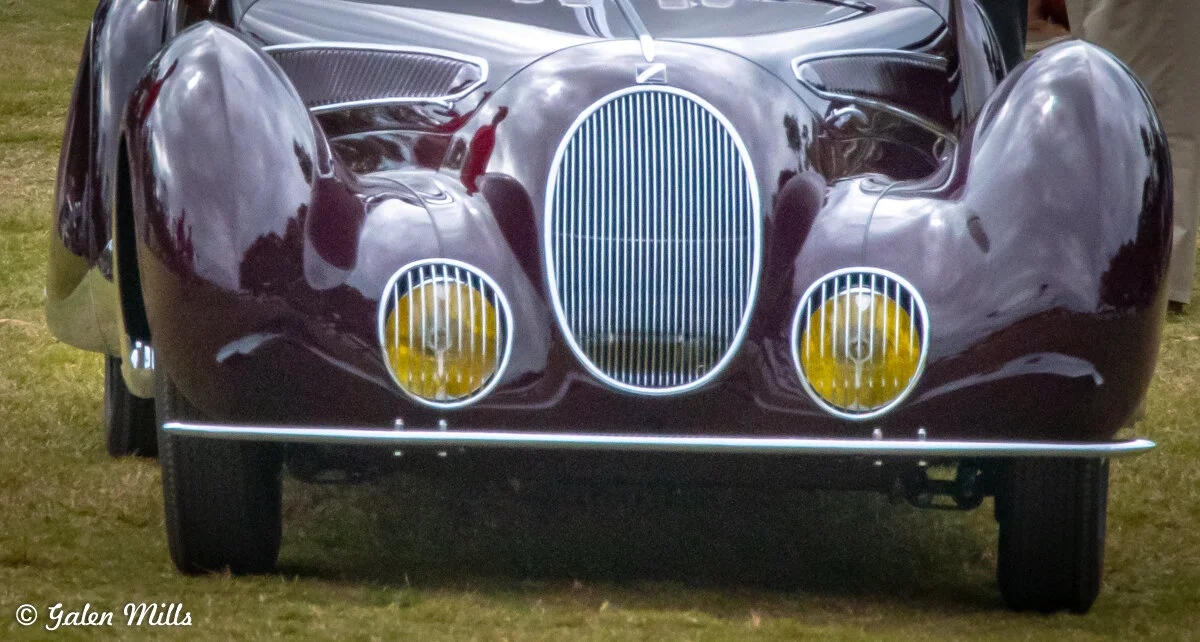 Front view of a vintage car with a glossy dark finish, featuring a prominent vertical grille and two yellow headlights, parked on grassy ground.