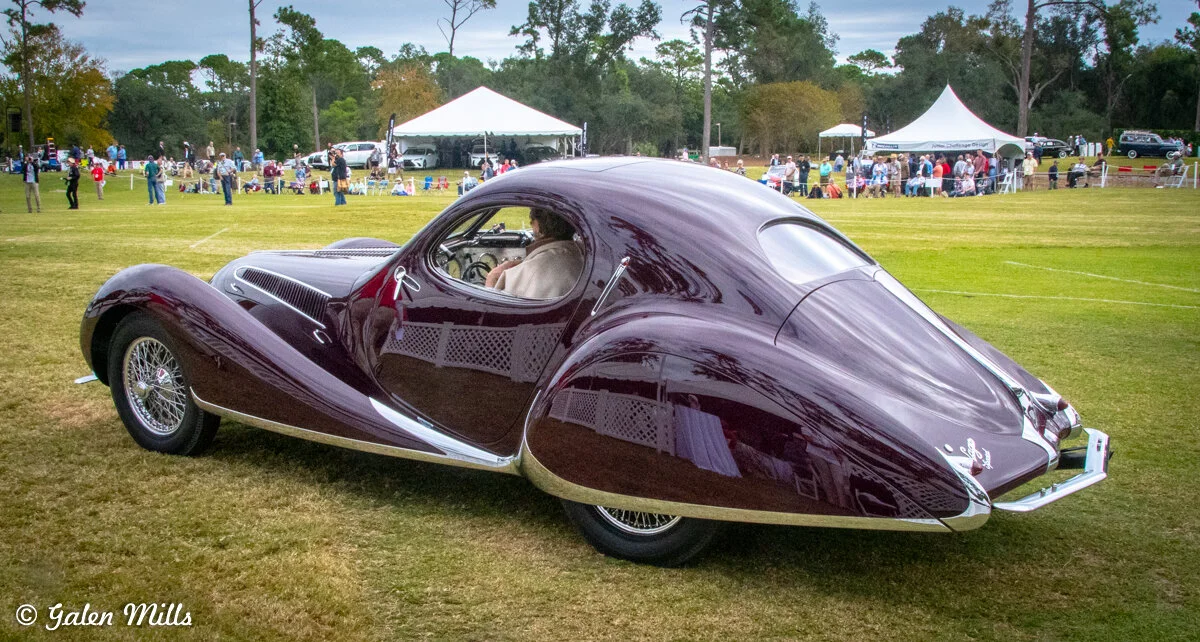Vintage purple classic car with curved design at an outdoor auto show.
