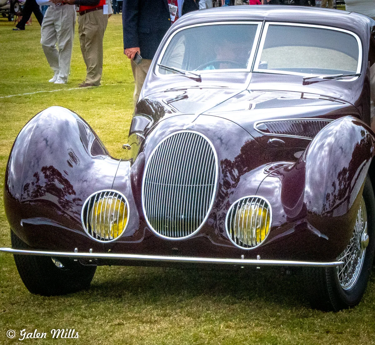 A vintage classic car with a maroon finish, showcasing an art deco design style with large rounded fenders, a prominent vertical grille, and yellow-tinted headlights, displayed on a grassy area.
