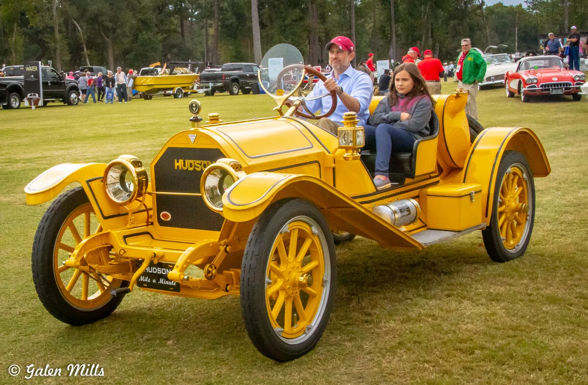 Vintage yellow Hudson automobile with two people seated, set in a grassy area among other classic cars, including a red convertible in the background.