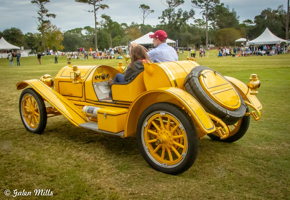 A vintage yellow car with two passengers on a grassy field, surrounded by event tents and people in the background.