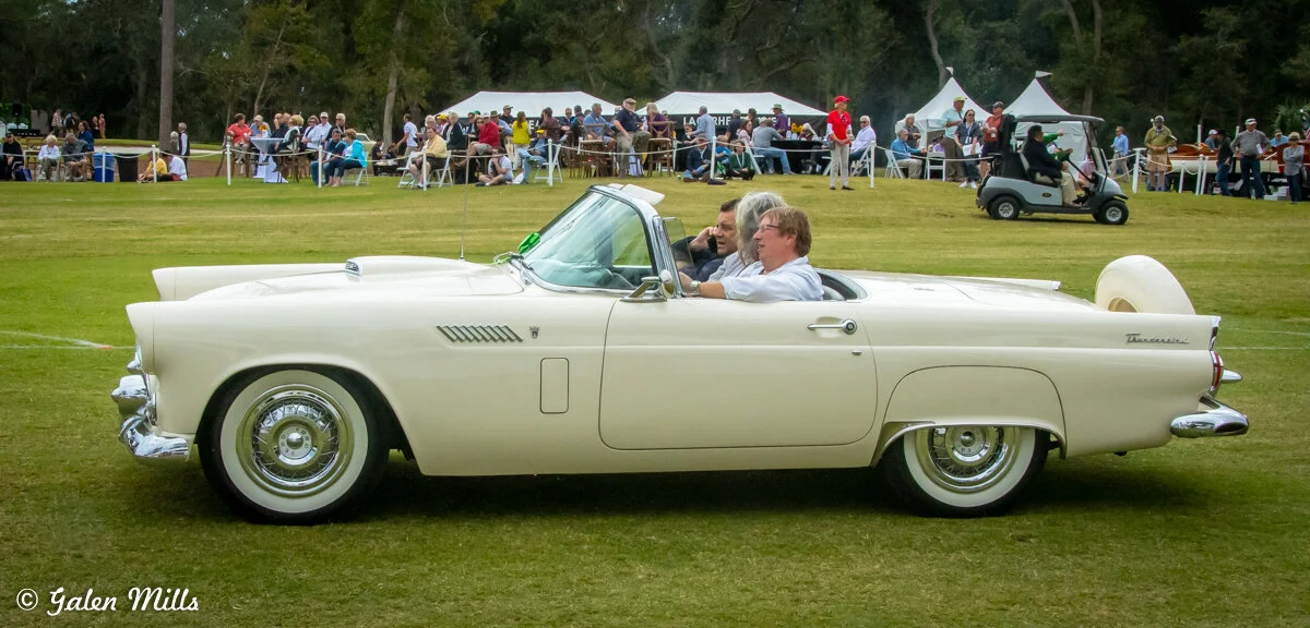 Vintage white convertible car with two passengers at an outdoor event with spectators and tents in the background.