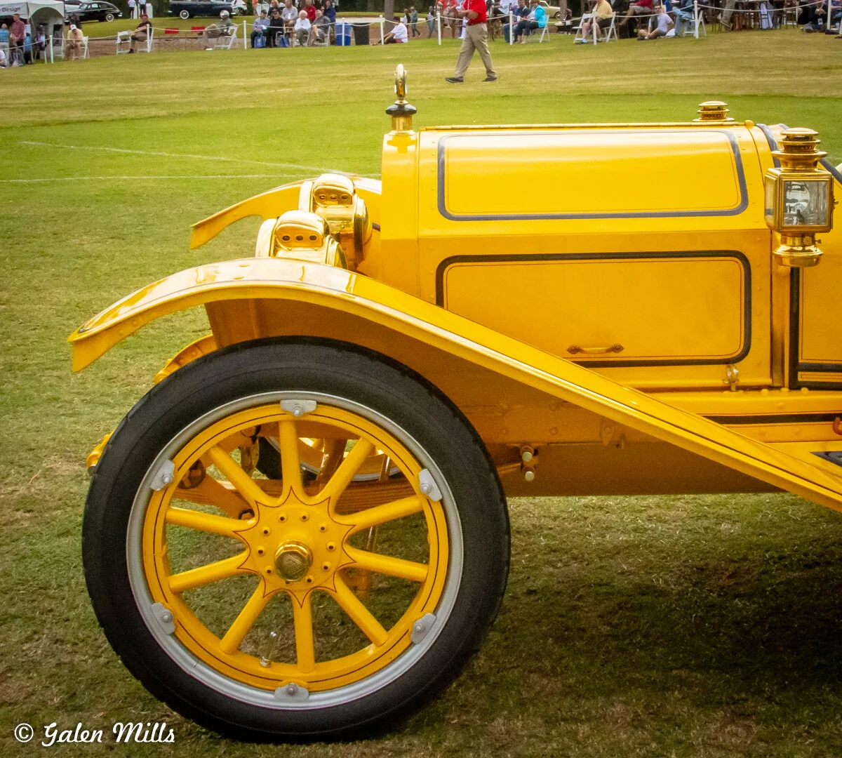 Yellow vintage car with intricate details on grass, showing front wheel and part of the body, with people in the background.