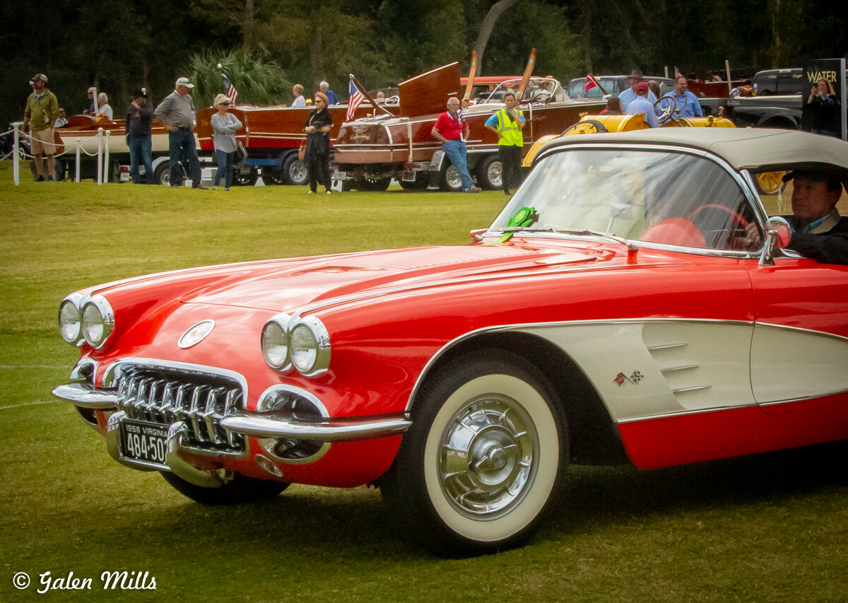 Vintage red and white convertible car on display at an outdoor event, with people and classic boats in the background.