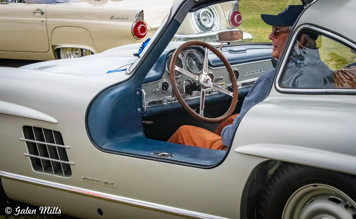 Person sitting in a classic Mercedes-Benz 300SL Gullwing with an open door, vintage cars in the background.