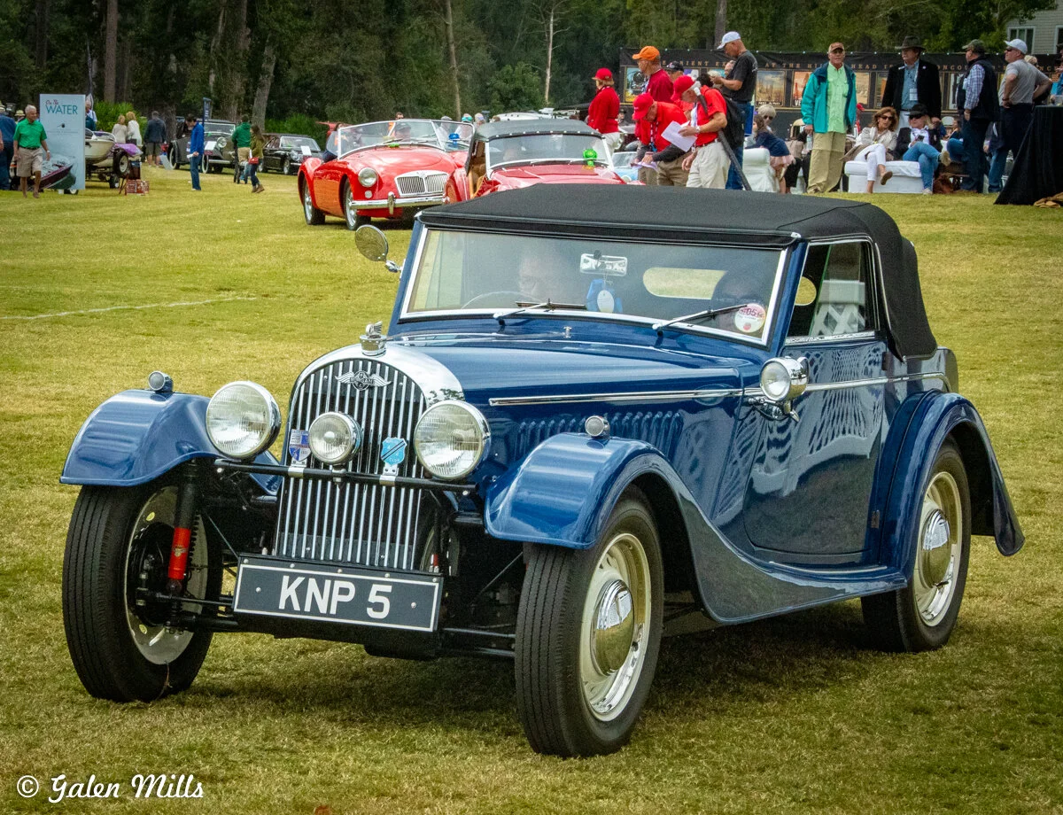 Classic blue Morgan car at a car show with other vintage cars and people in the background.