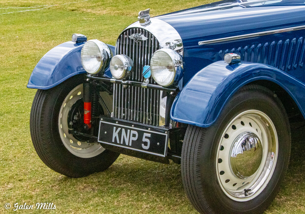Front view of a vintage blue Morgan car with two large circular headlights and a grill, parked on grass.