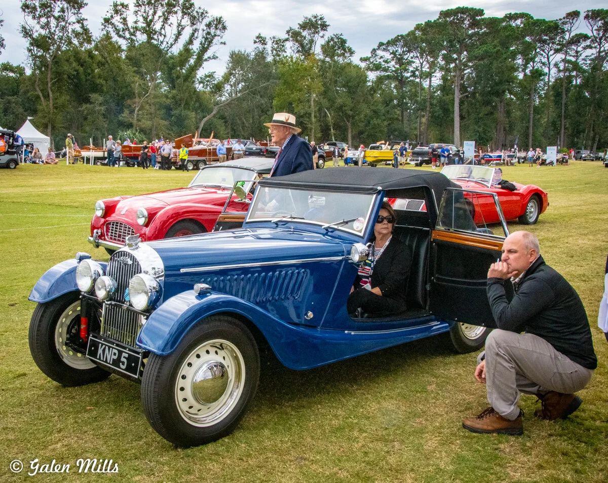 Vintage car show with a classic blue convertible in the foreground and people gathered around, surrounded by trees and more vintage vehicles in a park setting.