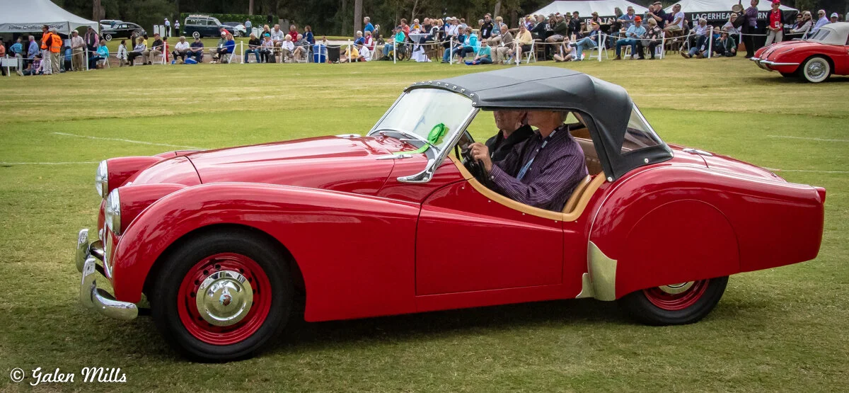 A vintage red convertible car with a black soft top, parked on grass, surrounded by spectators at an outdoor car show.