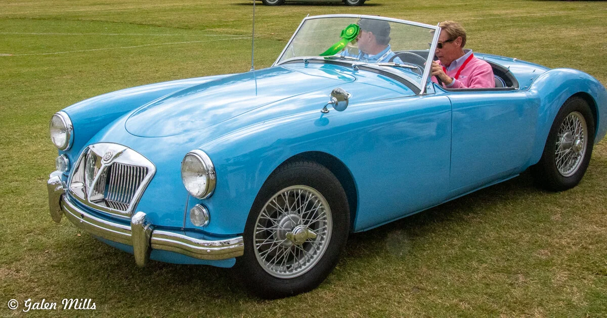 Classic blue MG car at a car show, with two people inside, one wearing sunglasses, on a grassy field.