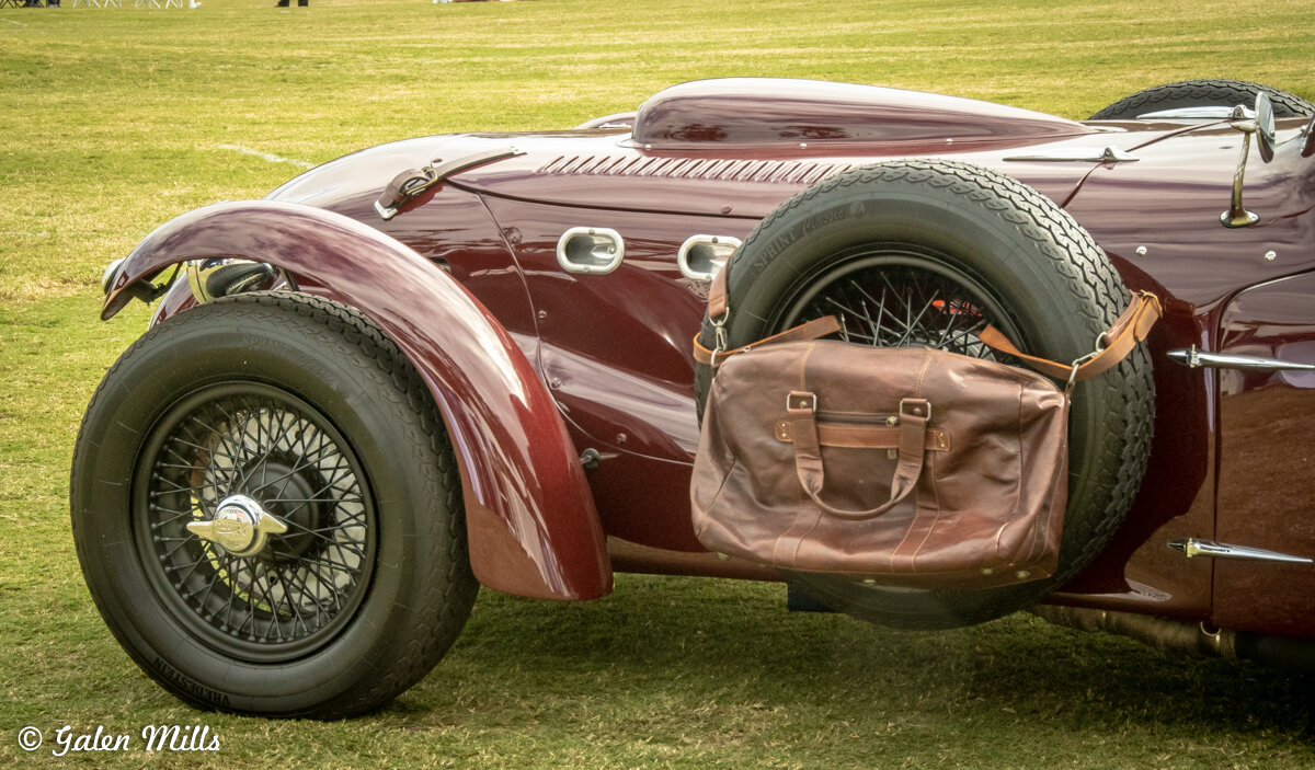 Vintage maroon car with spare tire and brown leather duffel bag on grass