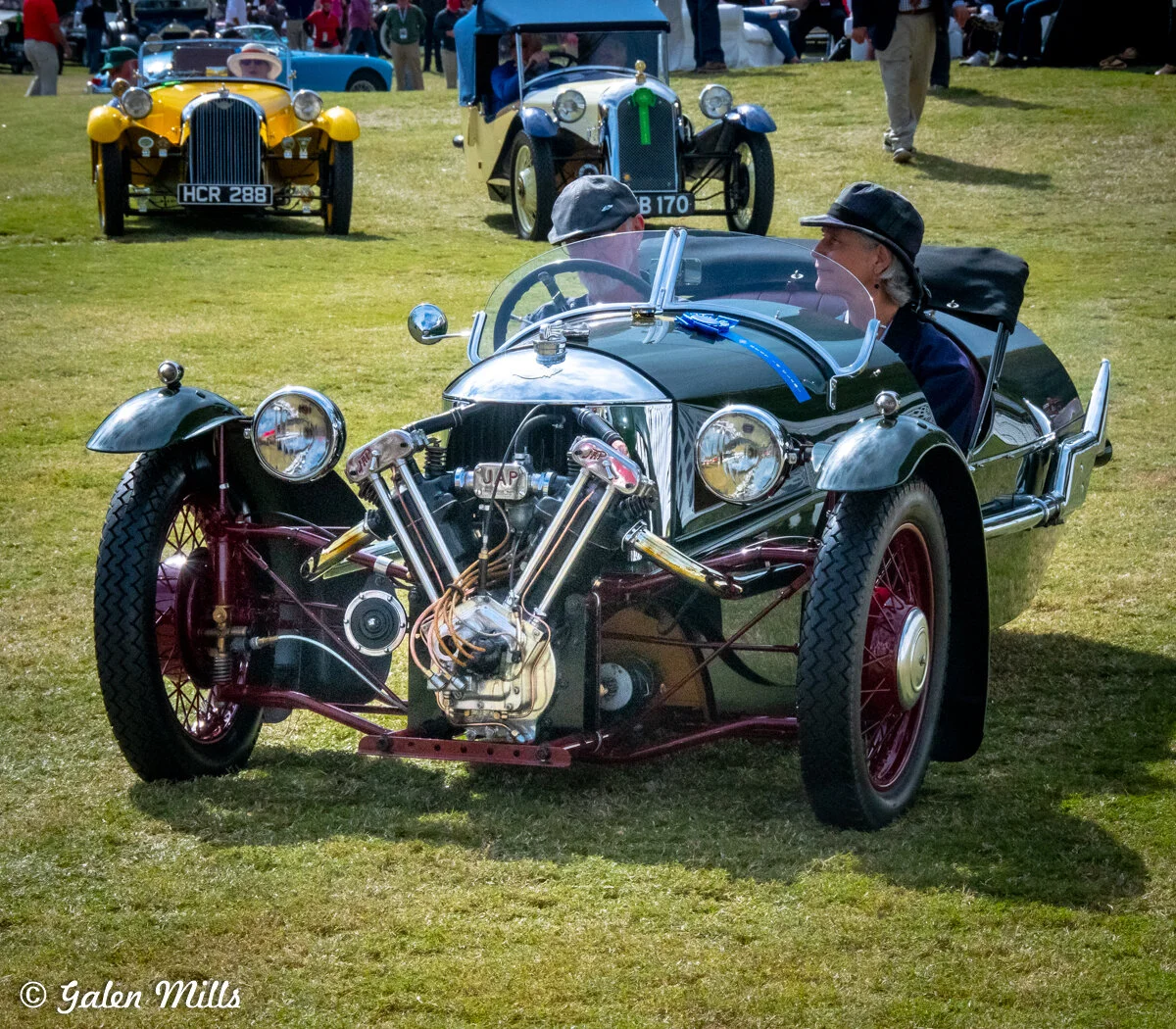 Vintage three-wheel car on grass with other classic vehicles in background at a car show.