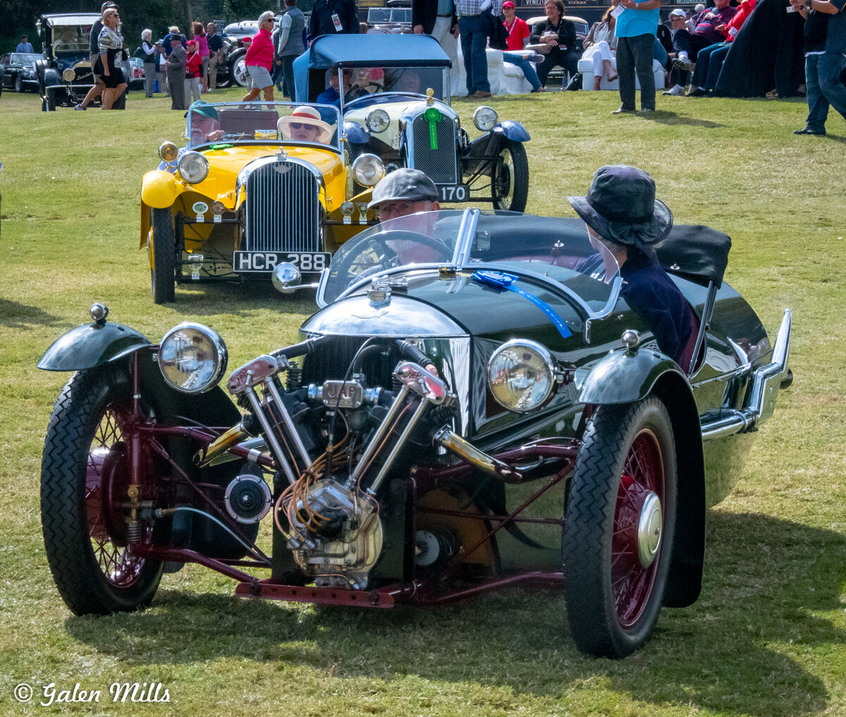Vintage cars displayed on grass with people observing at a car show.