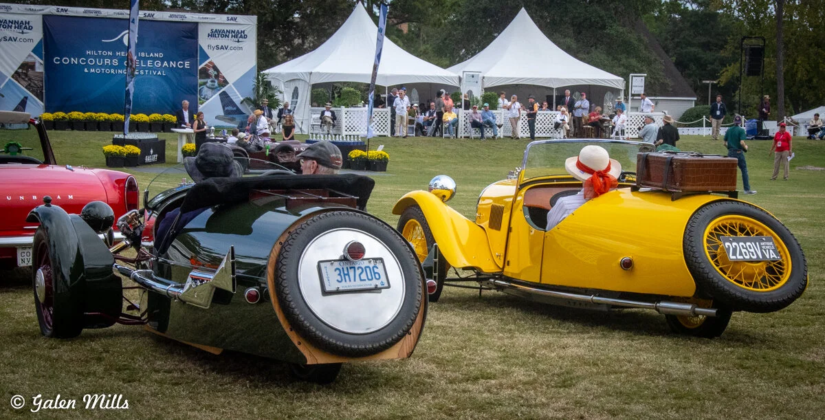 Vintage cars displayed at the Concours d'Elegance event with people in the background, featuring tents and a festival banner.