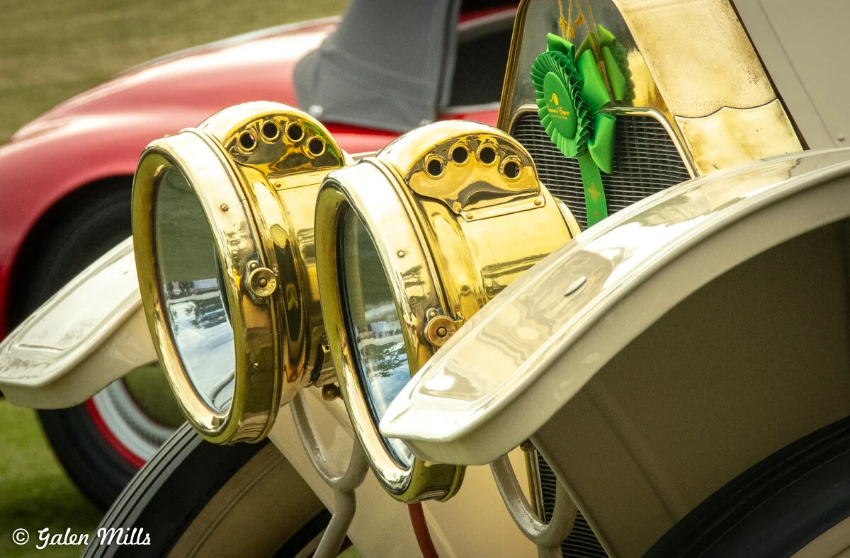 Close-up of vintage car headlights with brass detailing and a green award ribbon on the grill; part of a classic car show.