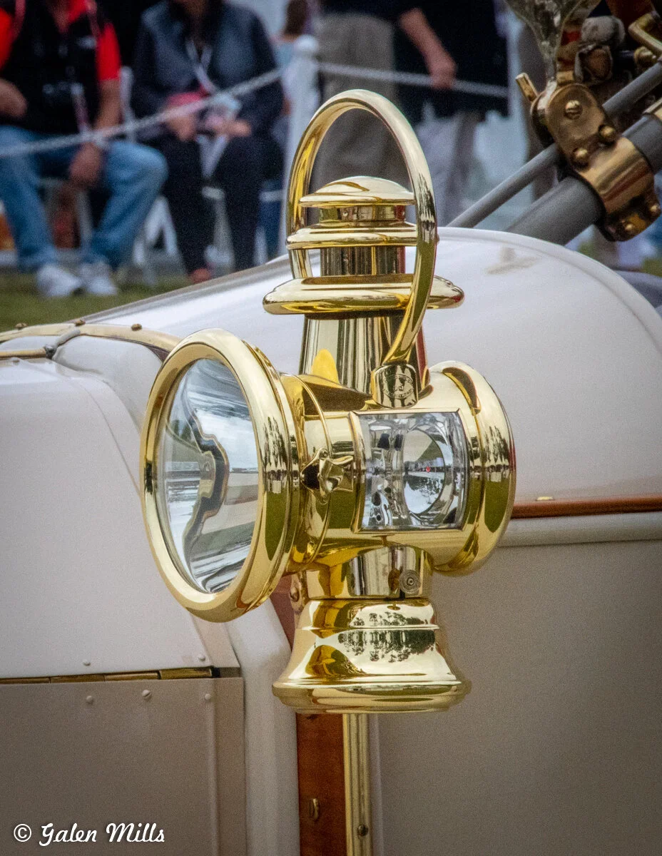 Close-up of a vintage car with a brass headlamp and decorative details, with blurred people in the background.