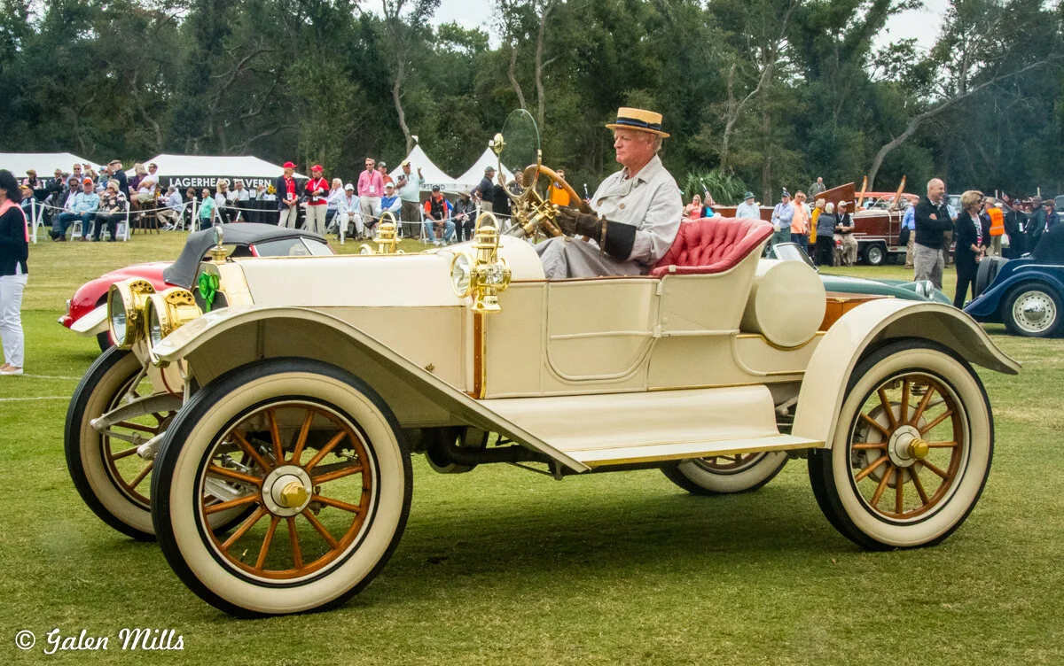 A vintage car with a man in period attire driving it at an outdoor event, surrounded by spectators. The car is cream-colored with red seats and brass accents.