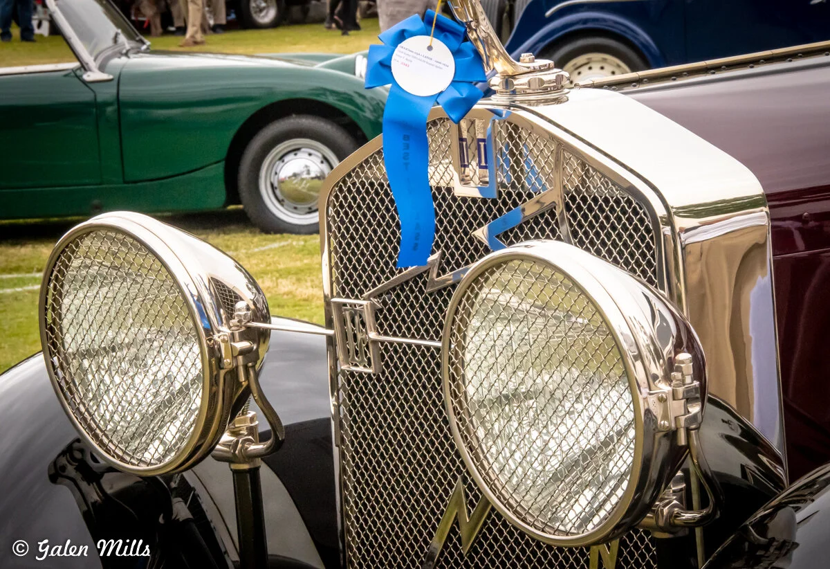 Close-up of a vintage car with a blue ribbon award on the grille, showcasing chrome headlights and a classic design. Other classic cars are visible in the background.