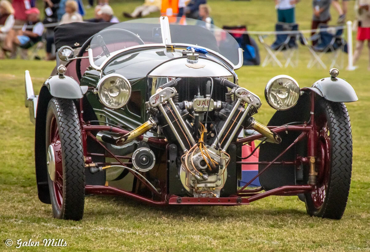 Front view of a vintage Morgan three-wheeler car with an exposed engine, large headlights, and wire-spoke wheels, parked on grass with people in the background.