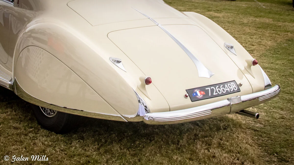Rear view of a vintage car with a pale yellow exterior and chrome bumper, parked on grass.