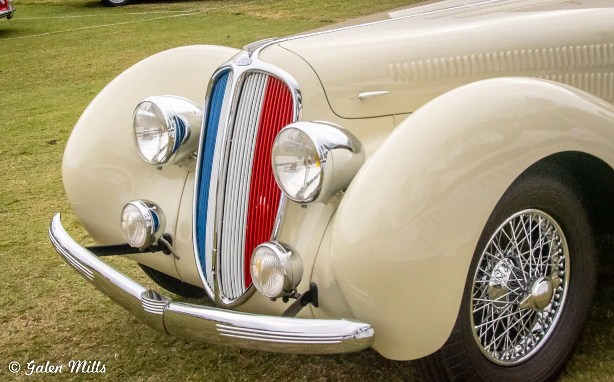 Front view of a vintage classic car featuring a red and blue vertical grille, chrome bumper, and wire-spoke wheel.
