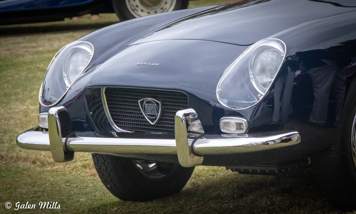Close-up of the front of a classic Lancia car, focusing on the grille, headlights, and chrome bumper, parked on grass.