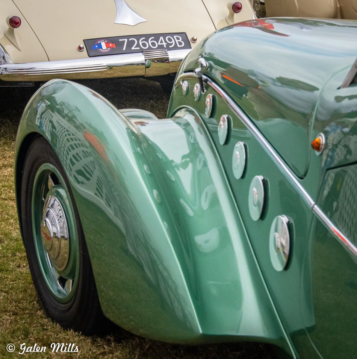 Close-up of two vintage cars, one green and one beige, with detailed views of their fenders and license plate areas. The green car has chrome details and a visible wheel, while the beige car's rear is noticeable in the background.
