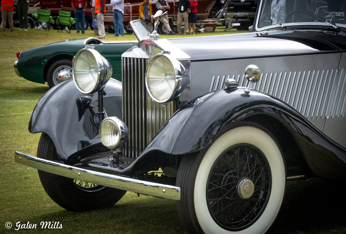 Vintage luxury car with classic design, featuring large headlights, a chrome grille, and white-wall tires, displayed at a car show on a grassy field.
