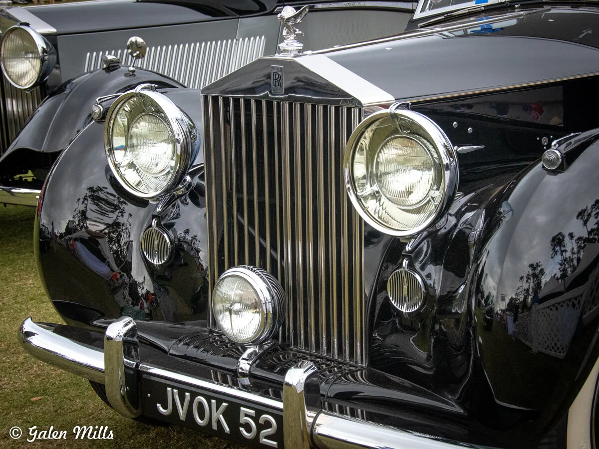 Close-up of the front of a classic black Rolls-Royce car with a chrome grille and round headlights at a car show.