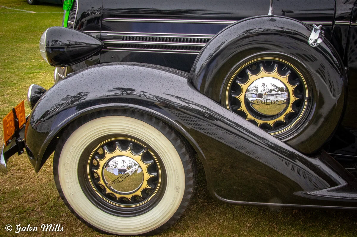 Close-up of an antique Dodge car with a focus on the wheel and hubcap, featuring a detailed design and reflective surface. The car has black body and white-walled tires, emphasizing vintage aesthetics.